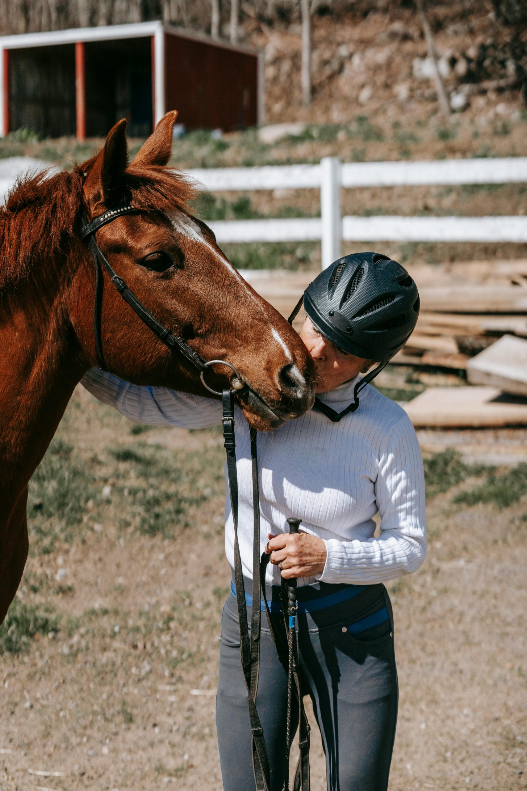 A woman wearing a riding helmet affectionately kisses a horse outdoors on a sunny day.